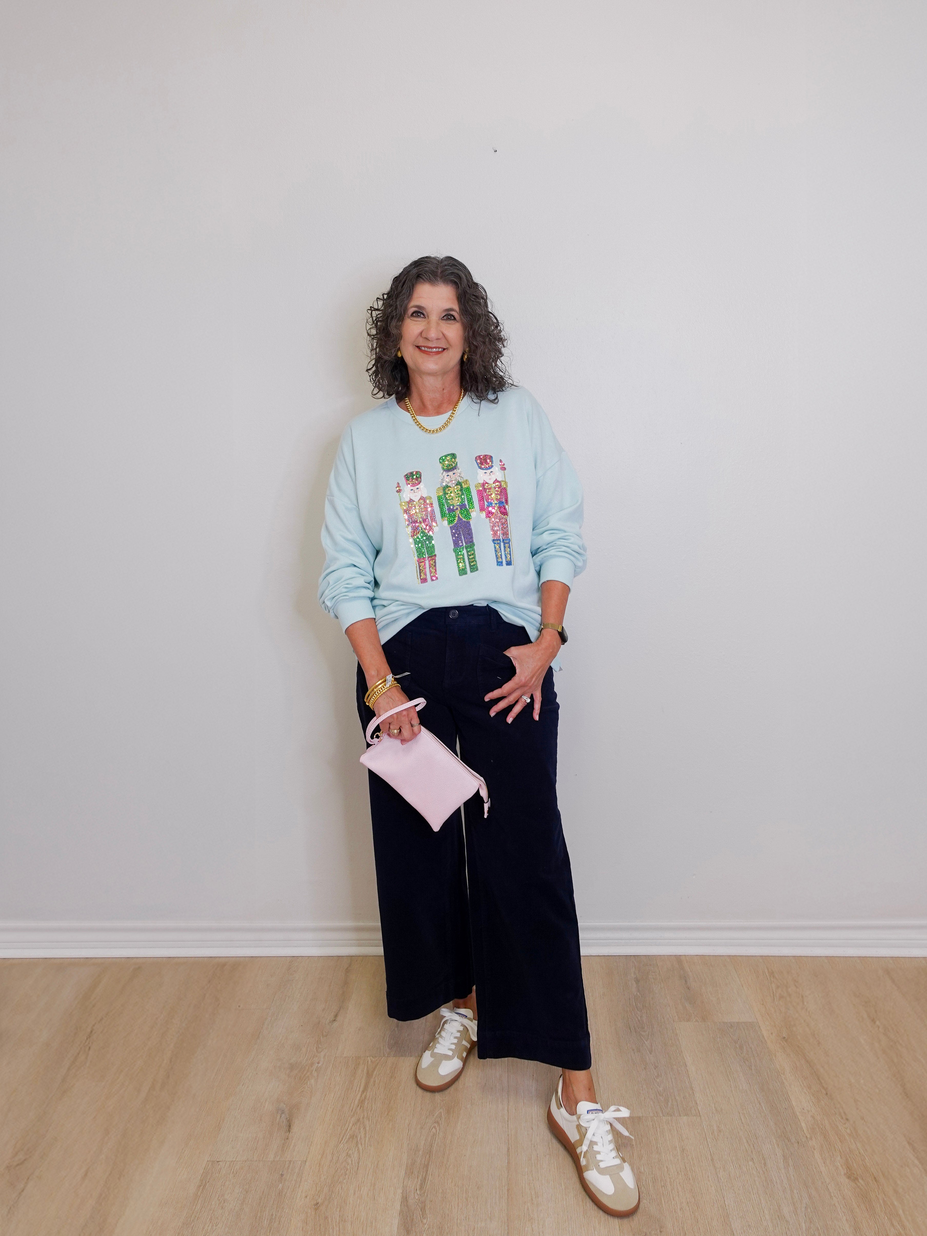 Woman wearing a light blue sweatshirt with a graphic design, dark pants, and white sneakers, holding a pink clutch.