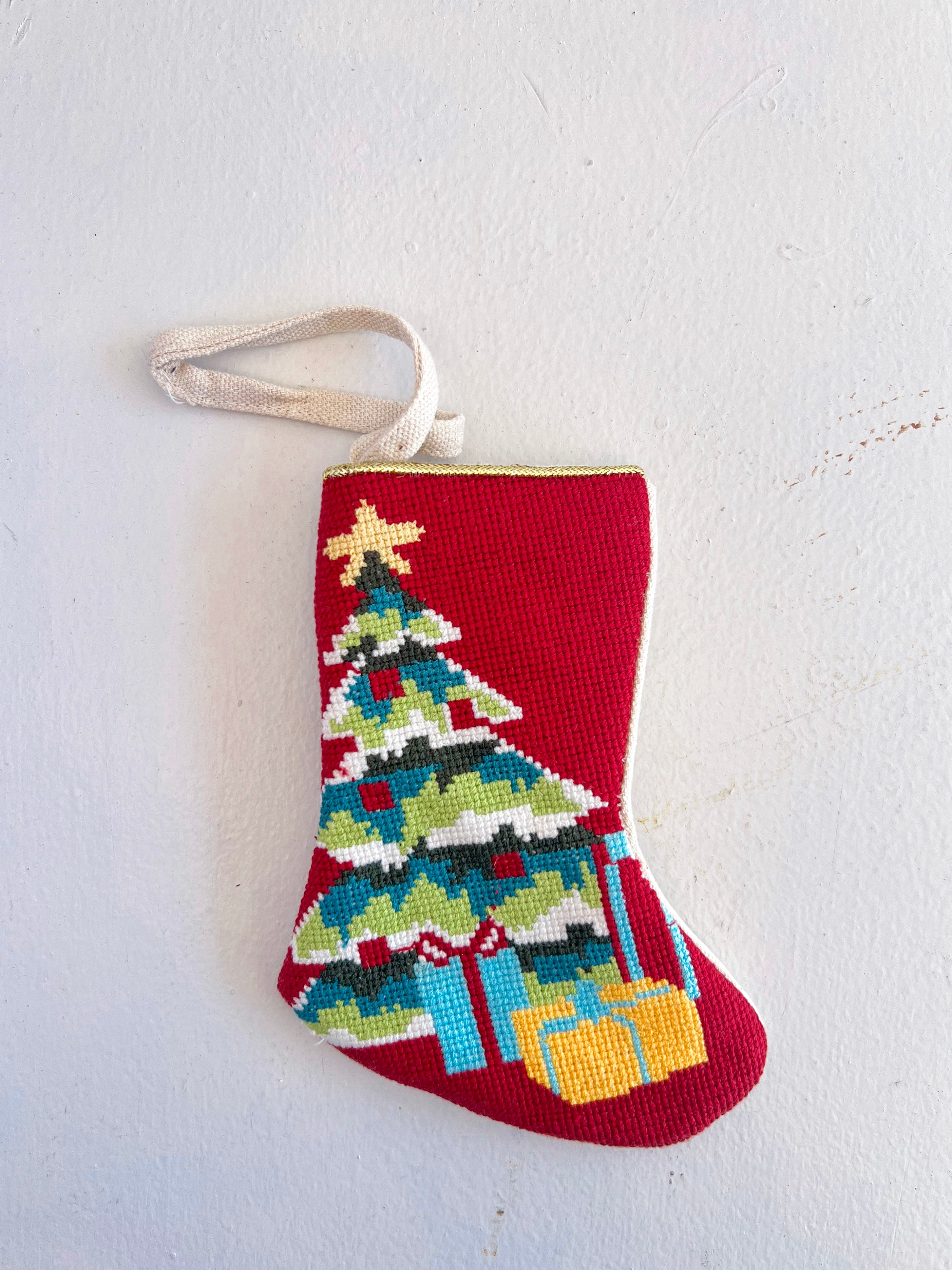 Red Christmas-themed needlepoint stocking with a tree and presents design on a white background