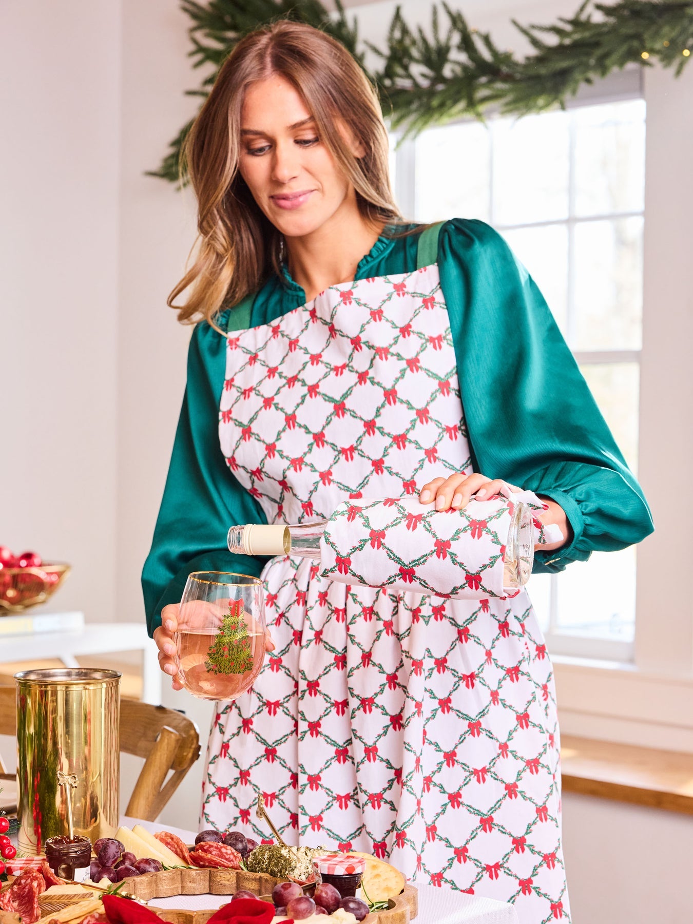 Woman in a kitchen wearing a patterned apron, holding a glass of wine and a bottle.