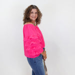 Woman wearing a bright pink blouse with ruffles and blue jeans, standing against a white wall.