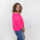 Woman wearing a bright pink blouse with ruffles and blue jeans, standing against a white wall.