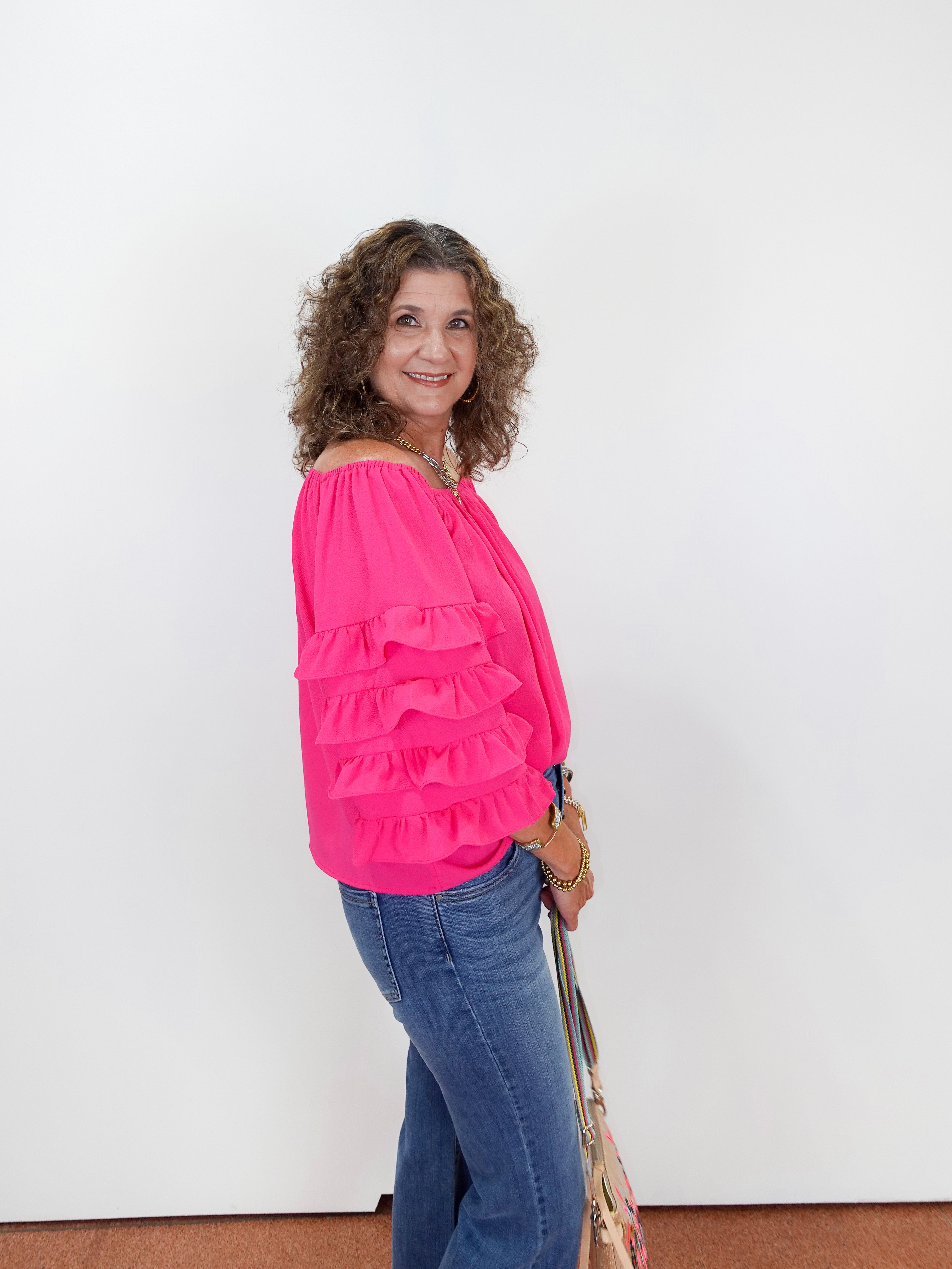 Woman wearing a bright pink blouse with ruffles and blue jeans, standing against a white wall.
