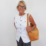 Woman wearing a white sleeveless top and patterned pants, holding a tan bag against a plain background