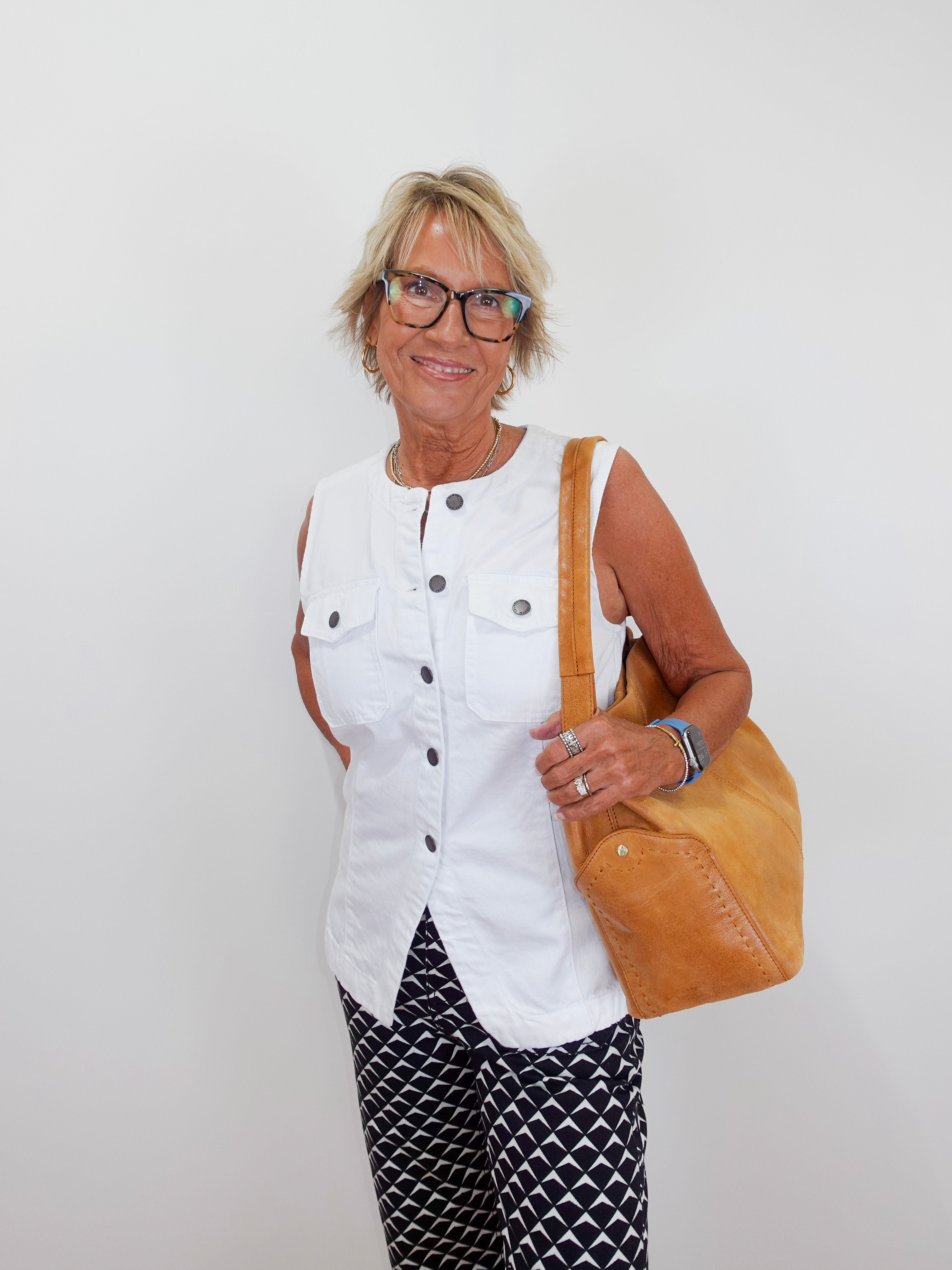 Woman wearing a white sleeveless top and patterned pants, holding a tan bag against a plain background