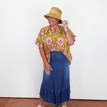 Woman wearing a patterned shirt, blue skirt, and straw hat on a plain background