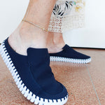 A person wearing dark navy slip-on mule shoes with white contrast stitching around the edges and a white sole, shown standing on a terrazzo floor.