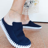A person wearing dark navy slip-on mule shoes with white contrast stitching around the edges and a white sole, shown standing on a terrazzo floor.