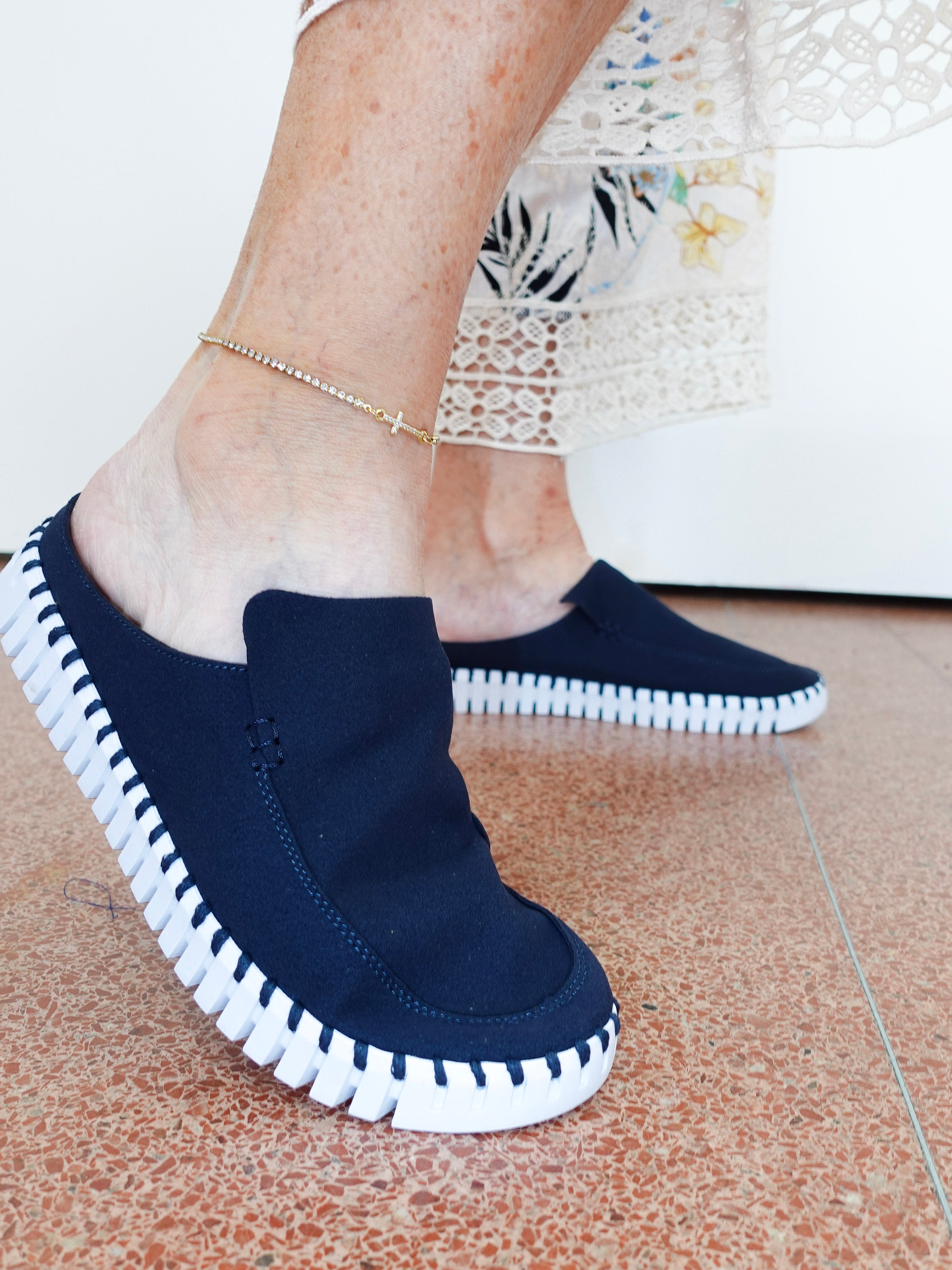A person wearing dark navy slip-on mule shoes with white contrast stitching around the edges and a white sole, shown standing on a terrazzo floor.
