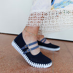 A person wearing dark navy blue knit Mary Jane-style slip-on shoes with two blue straps and gold-tone buckle details, paired with a white lace-trimmed dress and standing on a terracotta tiled floor.
