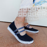 A person wearing dark navy blue knit Mary Jane-style slip-on shoes with two blue straps and gold-tone buckle details, paired with a white lace-trimmed dress and standing on a terracotta tiled floor.