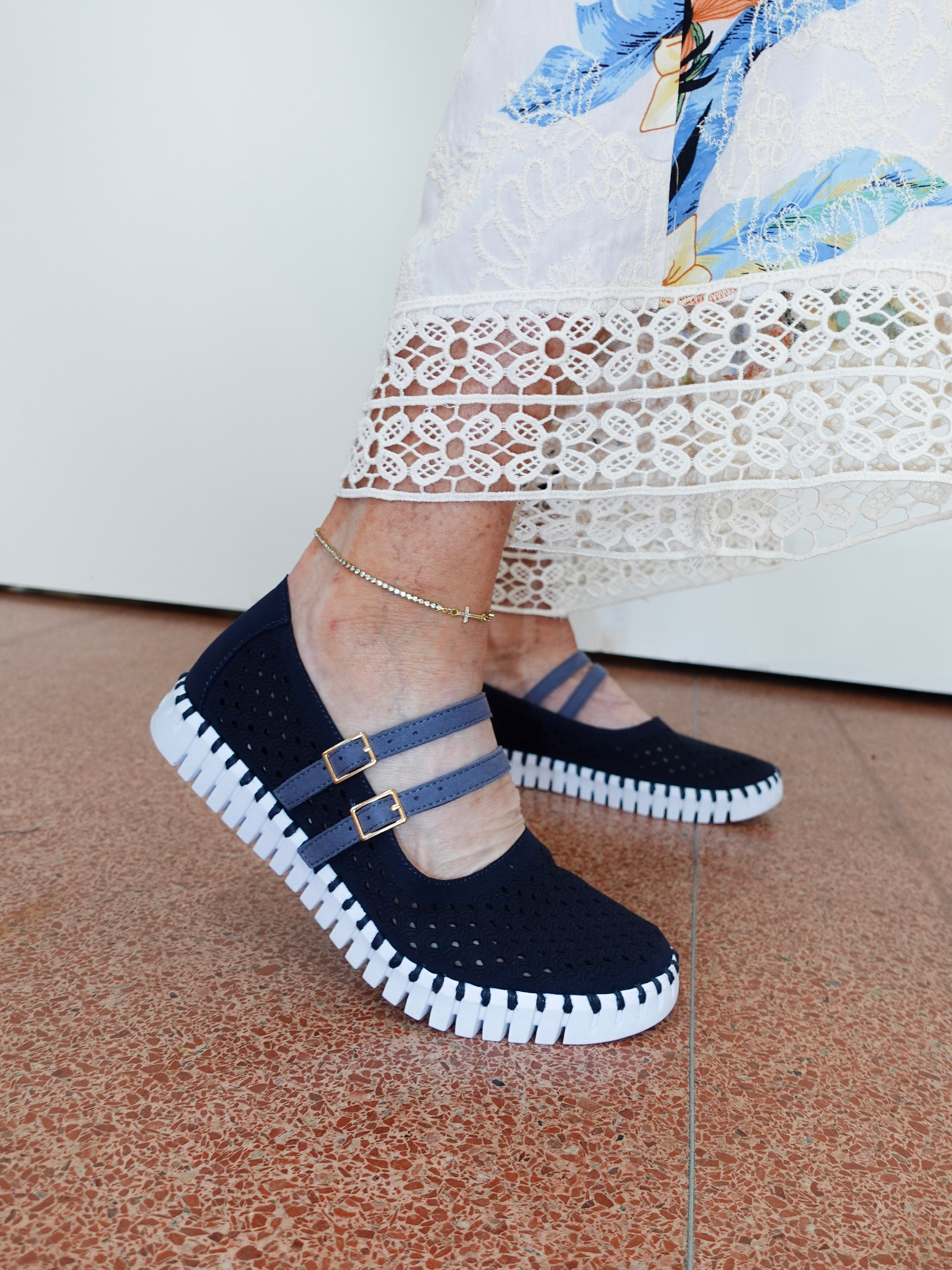 A person wearing dark navy blue knit Mary Jane-style slip-on shoes with two blue straps and gold-tone buckle details, paired with a white lace-trimmed dress and standing on a terracotta tiled floor.