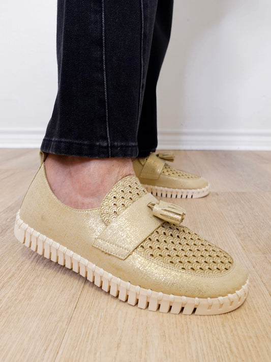 A person modeling a the ilse Jacobsen tassel loafer in gold against a white wall.