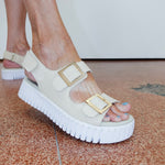 Beige sandals with gold buckles on a terrazzo floor.