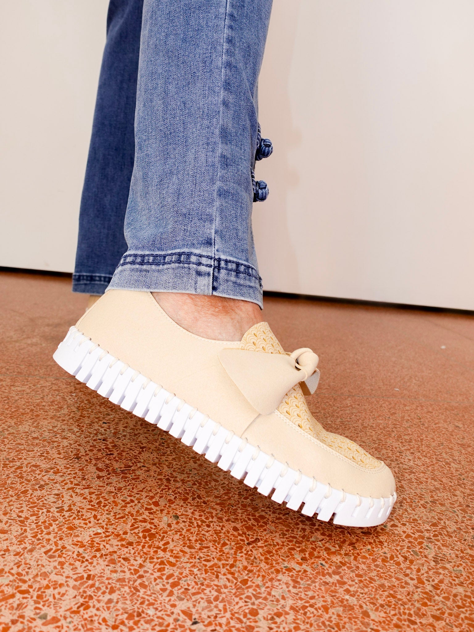 Beige sneakers with white soles worn with blue jeans on a speckled floor.