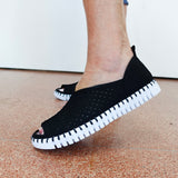 A person wearing black open-toe slip-on shoes with a textured black upper and a thick white ridged sole, standing on a speckled terracotta floor against a white wall.