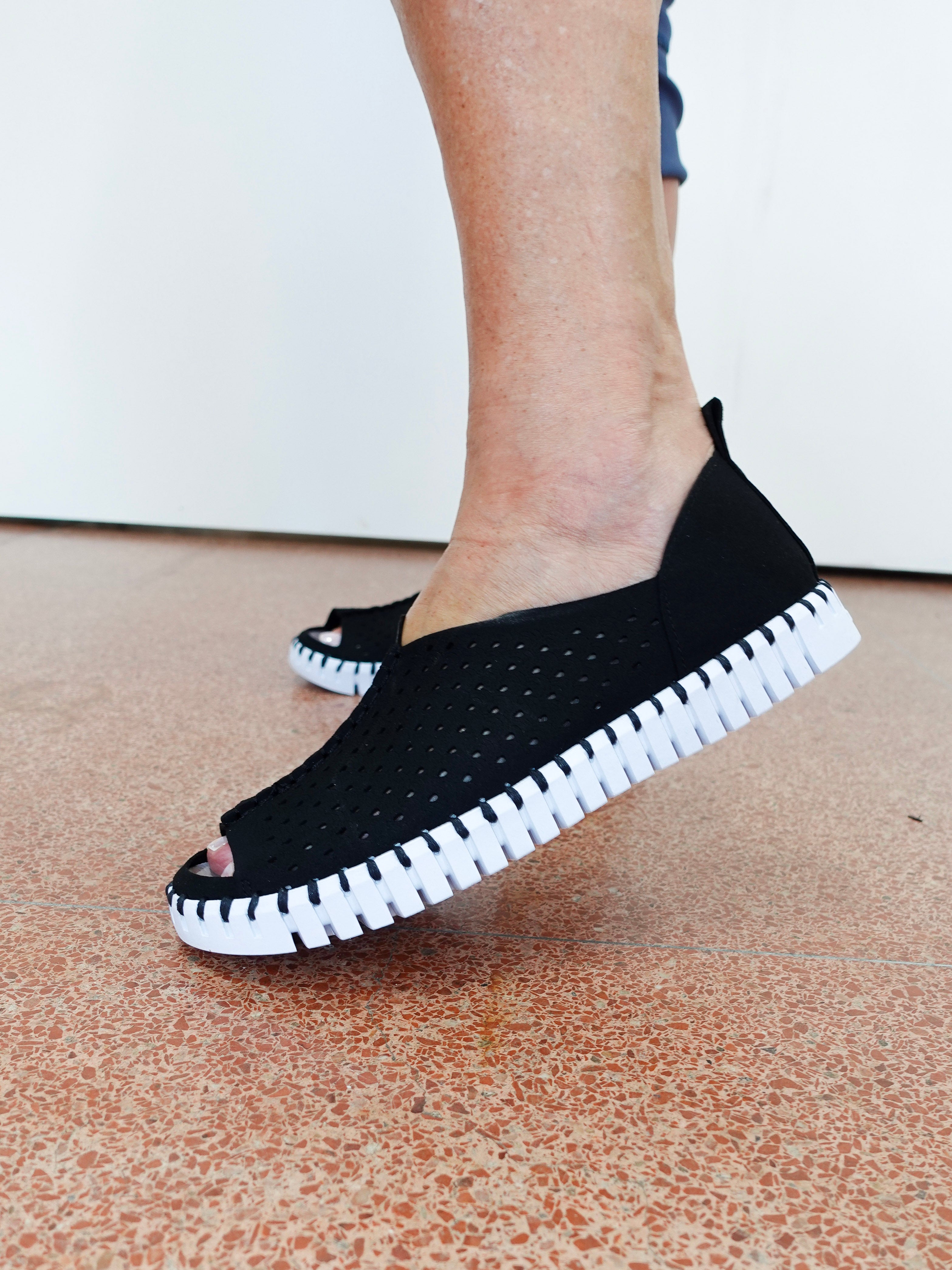 A person wearing black open-toe slip-on shoes with a textured black upper and a thick white ridged sole, standing on a speckled terracotta floor against a white wall.
