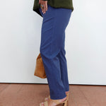 A person shown from the waist down wearing blue straight-leg trousers, an olive green knit sweater, beige heeled sandals, and holding a tan handbag against a white wall.
