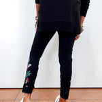 A person shown from the back wearing black slim-fit pants with colorful floral embroidery near the ankle, a black long-sleeve top, and white slip-on shoes against a plain white wall and terracotta floor.