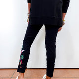 A person shown from the back wearing black slim-fit pants with colorful floral embroidery near the ankle, a black long-sleeve top, and white slip-on shoes against a plain white wall and terracotta floor.