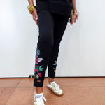 A person wearing black cropped pants with colorful floral embroidery on the lower legs, paired with a black sweatshirt and metallic gold sneakers, standing against a white wall.