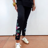 A person wearing black cropped pants with colorful floral embroidery on the lower legs, paired with a black sweatshirt and metallic gold sneakers, standing against a white wall.