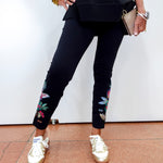 A person wearing black cropped pants with colorful floral embroidery near the cuffs, paired with shiny metallic gold sneakers, a black top, and holding a beige clutch against a plain white wall.