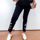 A person wearing black cropped pants with colorful floral embroidery near the cuffs, paired with shiny metallic gold sneakers, a black top, and holding a beige clutch against a plain white wall.