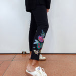 A person wearing black cropped pants with colorful floral embroidery on the lower leg, paired with white and gold sneakers, standing against a plain white wall.
