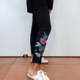 A person wearing black cropped pants with colorful floral embroidery on the lower leg, paired with white and gold sneakers, standing against a plain white wall.