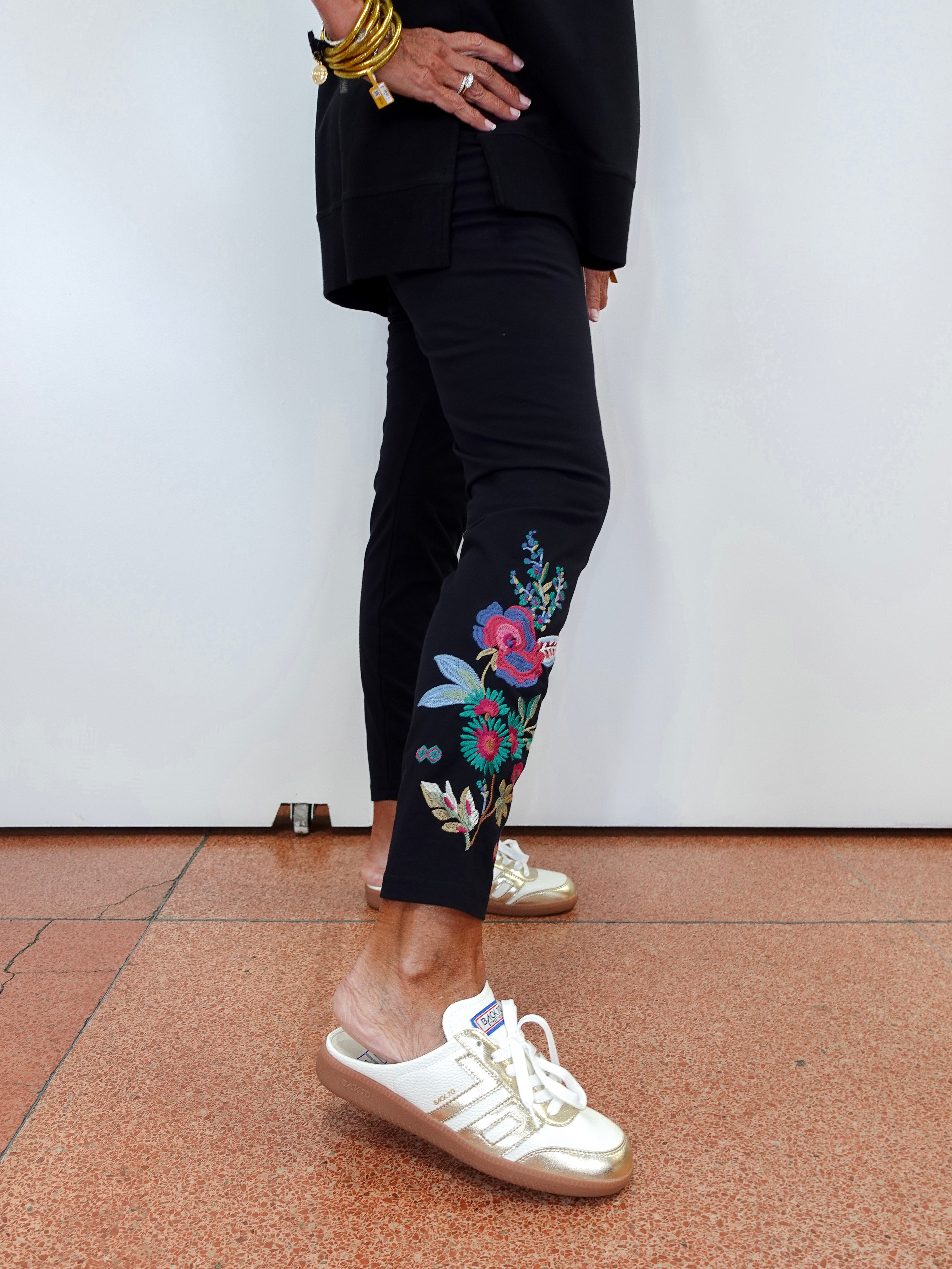 A person wearing black cropped pants with colorful floral embroidery on the lower leg, paired with white and gold sneakers, standing against a plain white wall.