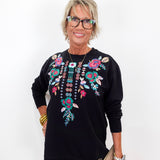A smiling older woman with short blonde hair and glasses wearing a black long-sleeve sweatshirt with colorful floral and bead embroidery on the chest, holding a beige clutch against a plain white background.
