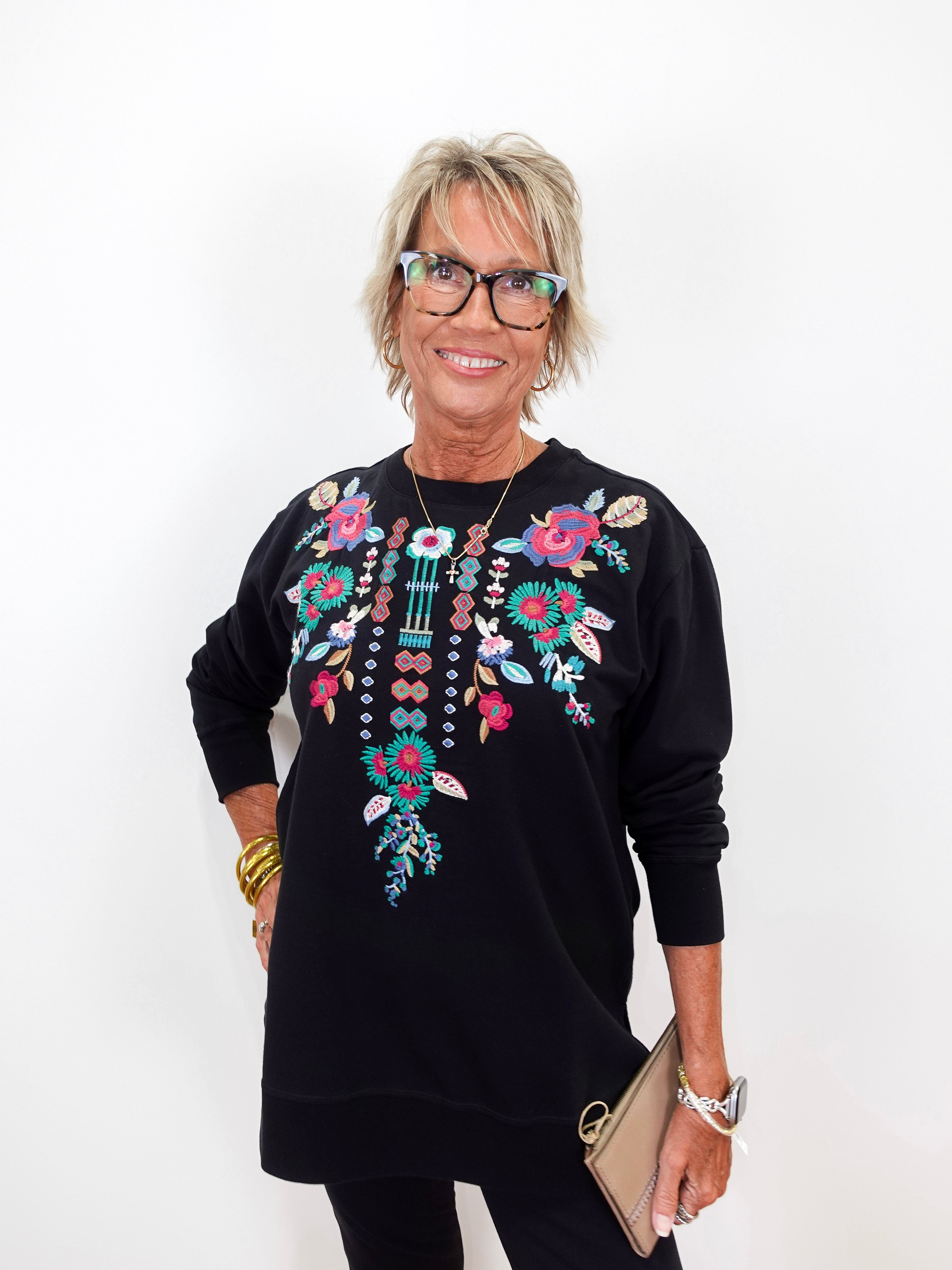A smiling older woman with short blonde hair and glasses wearing a black long-sleeve sweatshirt with colorful floral and bead embroidery on the chest, holding a beige clutch against a plain white background.