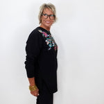 A woman with short gray hair and glasses, wearing a black long-sleeve top with colorful floral embroidery on the chest, standing against a plain white wall.