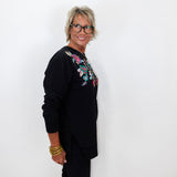 A woman with short gray hair and glasses, wearing a black long-sleeve top with colorful floral embroidery on the chest, standing against a plain white wall.
