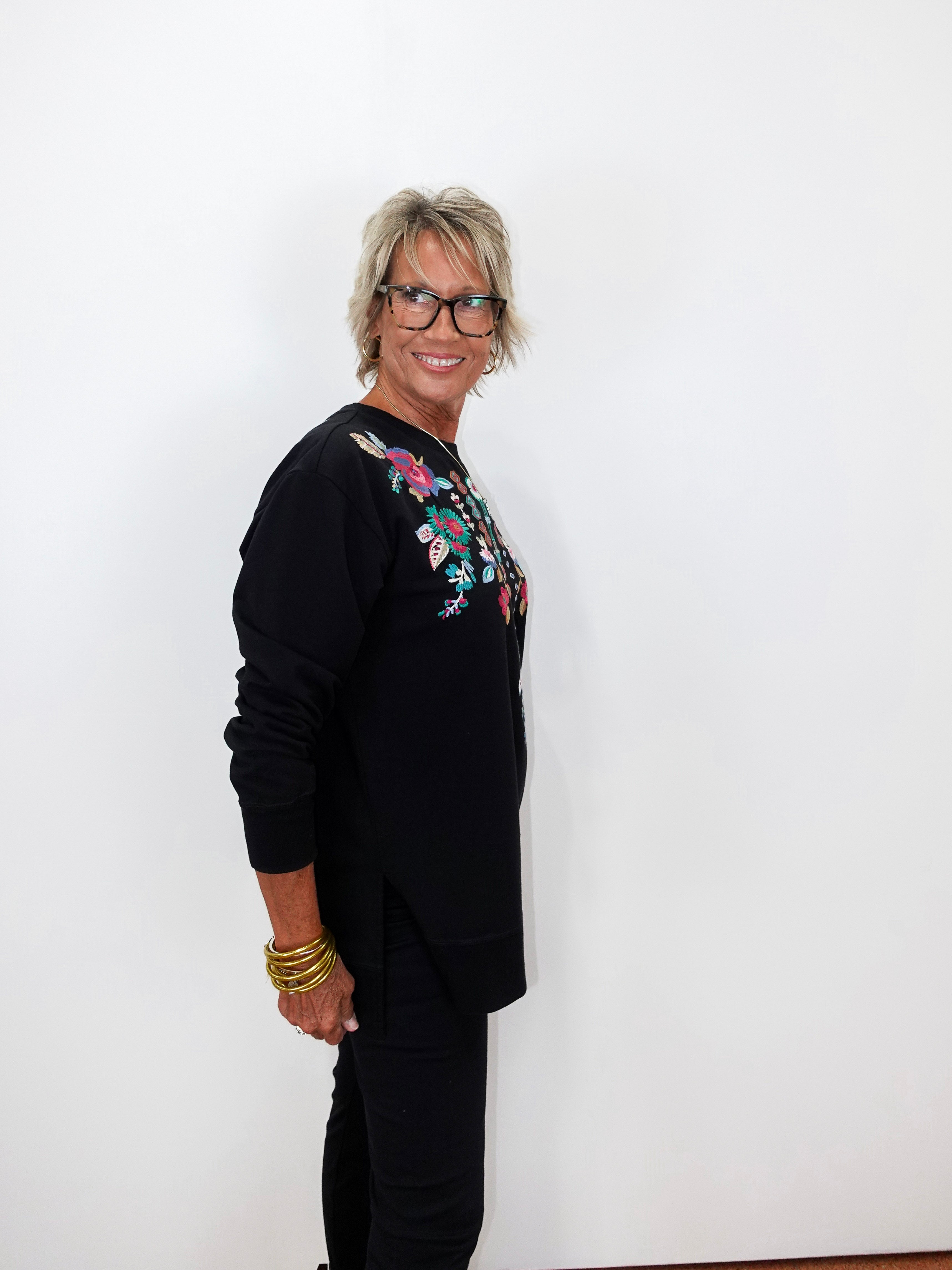 A woman with short gray hair and glasses, wearing a black long-sleeve top with colorful floral embroidery on the chest, standing against a plain white wall.