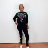 A woman with short blonde hair and glasses stands against a white wall wearing a black long-sleeve top with colorful floral embroidery on the chest, black pants with matching embroidery near the ankles, and white sneakers.