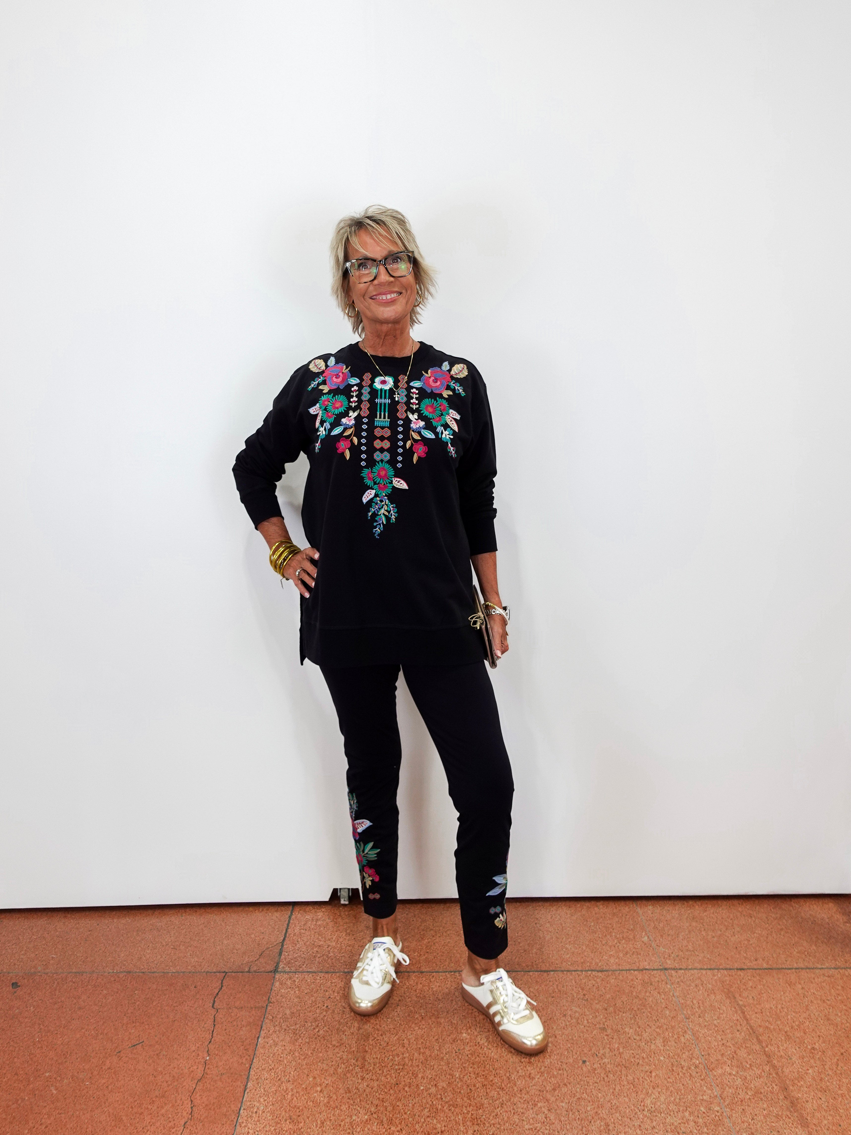 A woman with short blonde hair and glasses stands against a white wall wearing a black long-sleeve top with colorful floral embroidery on the chest, black pants with matching embroidery near the ankles, and white sneakers.