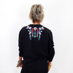 A person viewed from the back wearing a black long-sleeve sweatshirt with colorful floral embroidery across the upper back, paired with black pants against a plain white background.