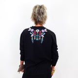 A person viewed from the back wearing a black long-sleeve sweatshirt with colorful floral embroidery across the upper back, paired with black pants against a plain white background.