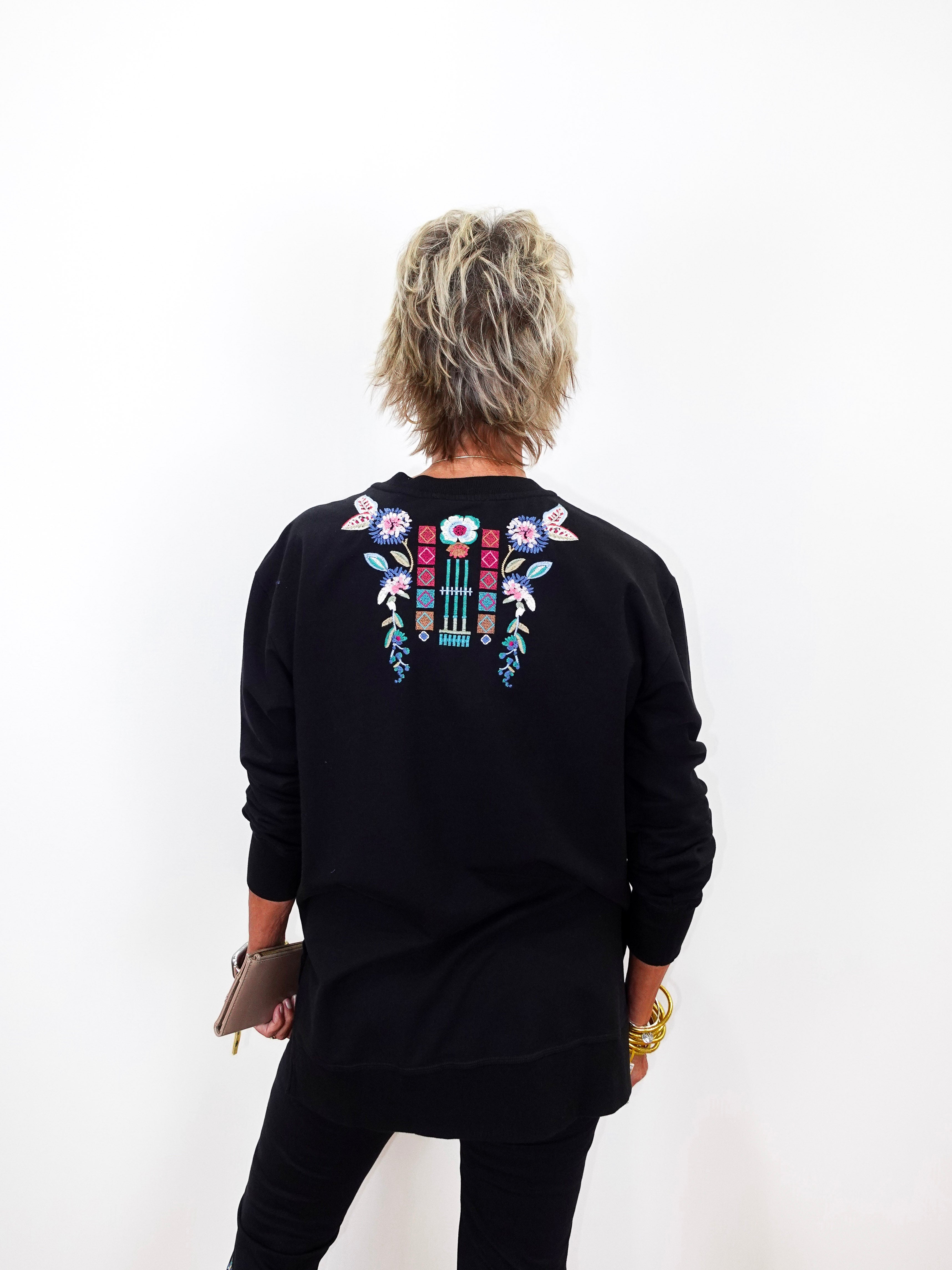 A person viewed from the back wearing a black long-sleeve sweatshirt with colorful floral embroidery across the upper back, paired with black pants against a plain white background.