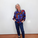 An older woman with gray hair stands against a plain white wall wearing a colorful abstract print button-up shirt in blue, red, and purple, paired with dark blue jeans and black loafers.