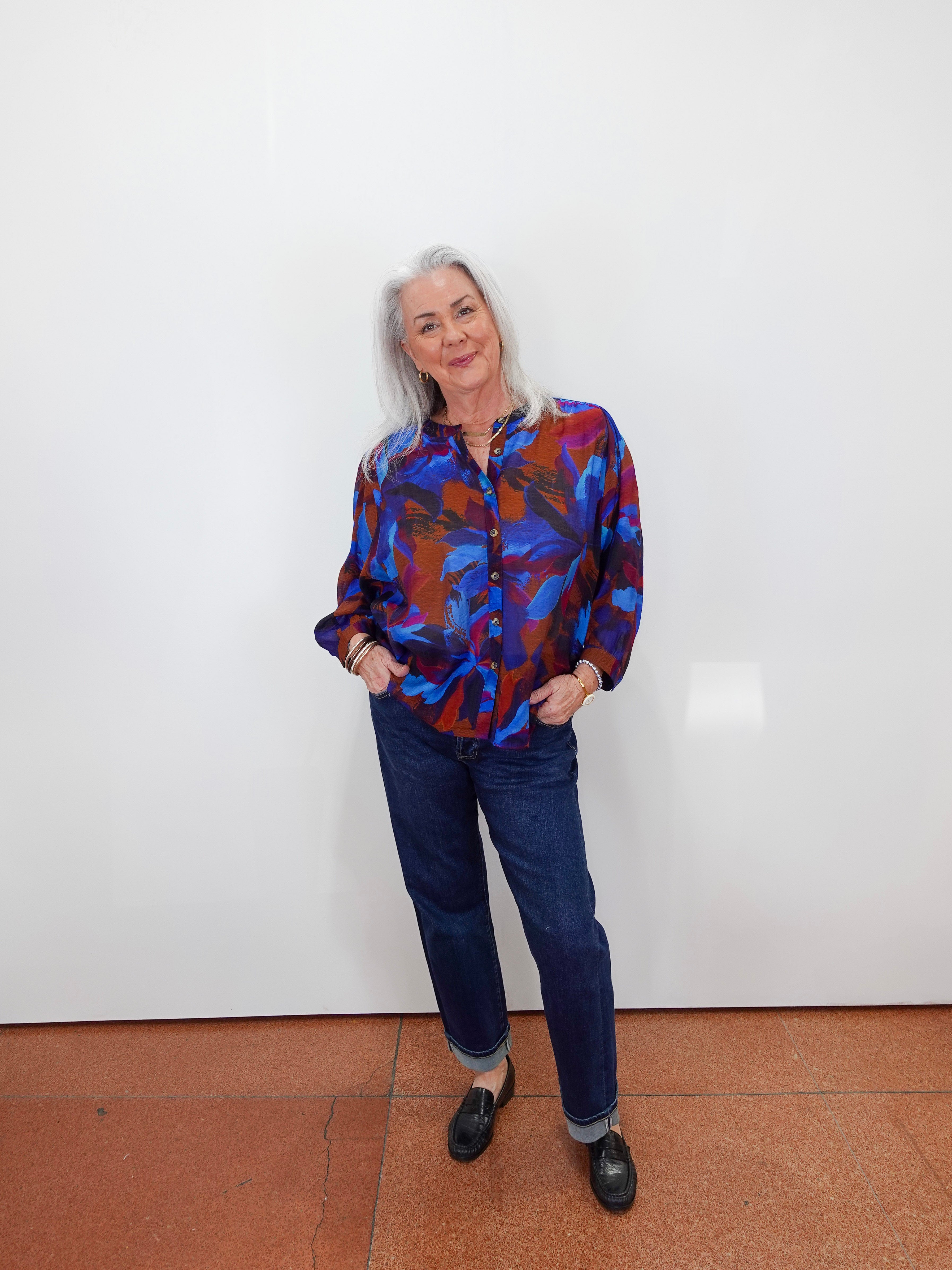 An older woman with gray hair stands against a plain white wall wearing a colorful abstract print button-up shirt in blue, red, and purple, paired with dark blue jeans and black loafers.