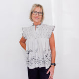 Woman wearing a light gray blouse with floral patterns and dark pants against a white background