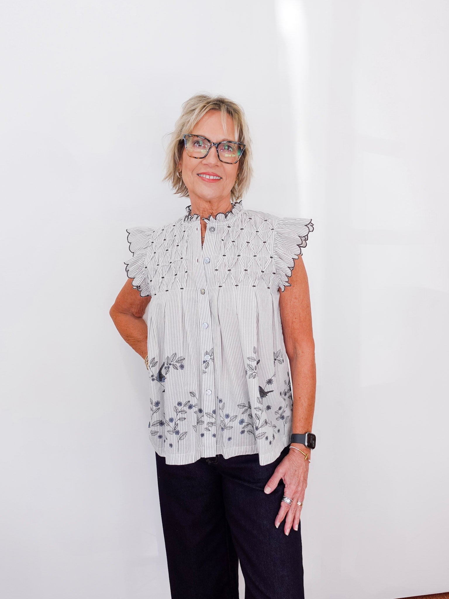 Woman wearing a light gray blouse with floral patterns and dark pants against a white background
