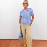 Woman wearing a blue patterned blouse and beige pants standing against a white wall.