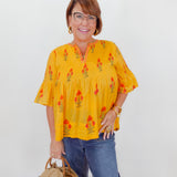 Woman wearing a yellow blouse with floral patterns and blue jeans, holding a straw bag against a white background.