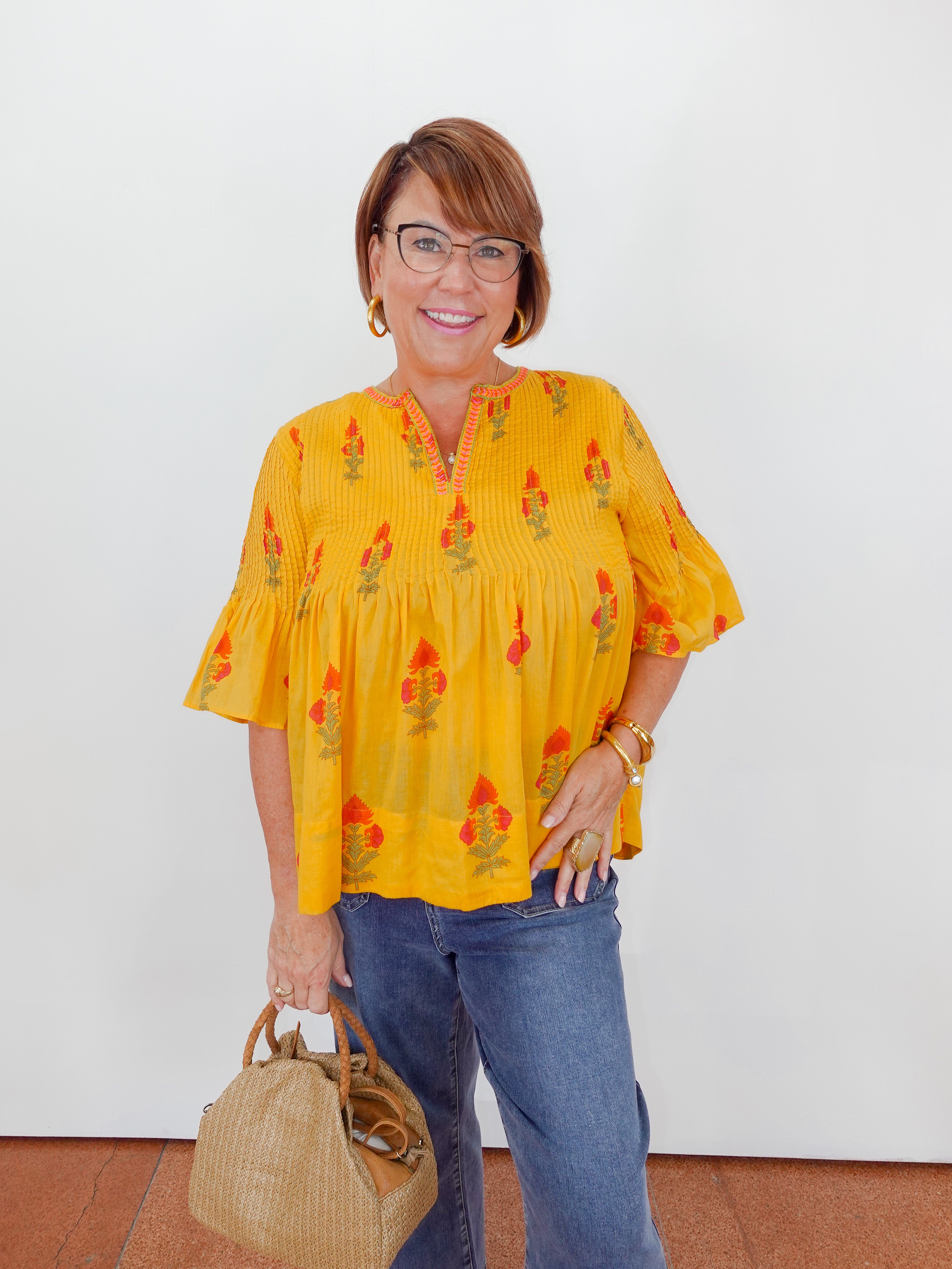 Woman wearing a yellow blouse with floral patterns and blue jeans, holding a straw bag against a white background.