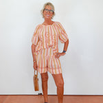 Woman wearing a colorful outfit with a striped skirt and top, holding a brown bag against a white wall.