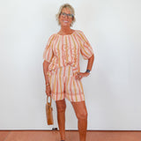 Woman wearing a colorful outfit with a striped skirt and top, holding a brown bag against a white wall.