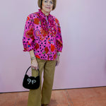 Woman wearing a colorful patterned blouse and holding a black handbag with a white bow against a pink wall.